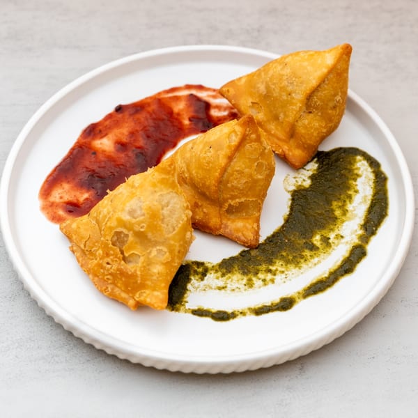 Three golden-brown Indian samosas on a white plate with red and green chutneys.